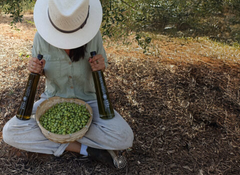 Teresa sitzt auf dem Boden des Olivenhains mit einem Korb voller Oliven und zwei Flaschen Isbilya Premium ungefiltertes natives Olivenöl extra.
