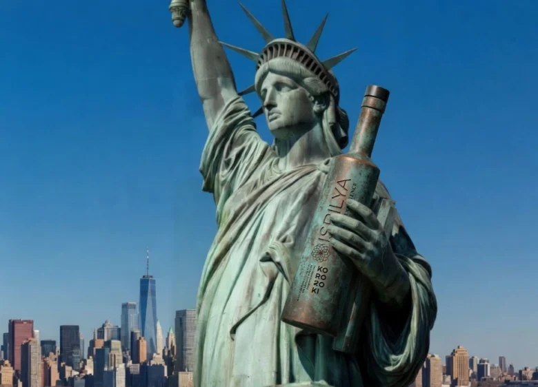 Primer plano de la Estatua de la Libertad abrazando una gran botella de bronce de aceite de oliva Isbilya Koroneiki con el skyline de Nueva York de fondo.